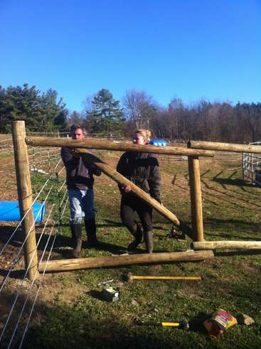 The image depicts two individuals constructing a fence, with one person holding a wooden beam and the other standing beside them, set against a backdrop of a field and trees.