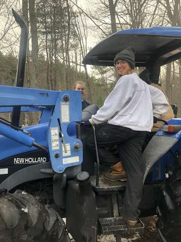 The image shows a woman sitting on a blue tractor, wearing a white sweatshirt, black pants, and brown boots, with a man standing behind her in a wooded area.
