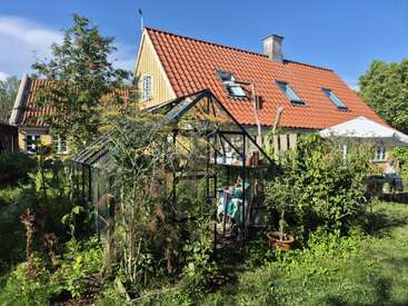 A sunny garden with a glass greenhouse, lush plants, and a yellow house with a red roof. Outdoor furniture and a person are visible.
