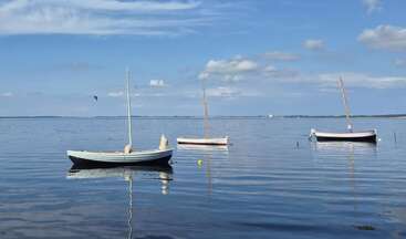 Three small boats float peacefully on calm, reflective water under a blue sky with scattered clouds. A bird soars above, adding life to the tranquil scene.