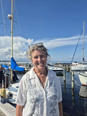 Uma mulher sorridente de cabelos grisalhos, vestindo uma blusa branca, está em frente a veleiros em uma marina sob um céu azul brilhante e parcialmente nublado.