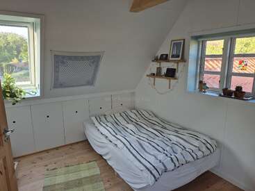 A cozy, minimalist bedroom features a striped bed, wooden floor, white walls, two windows, shelves with photos, potted plants, a wall hanging, and natural light.
