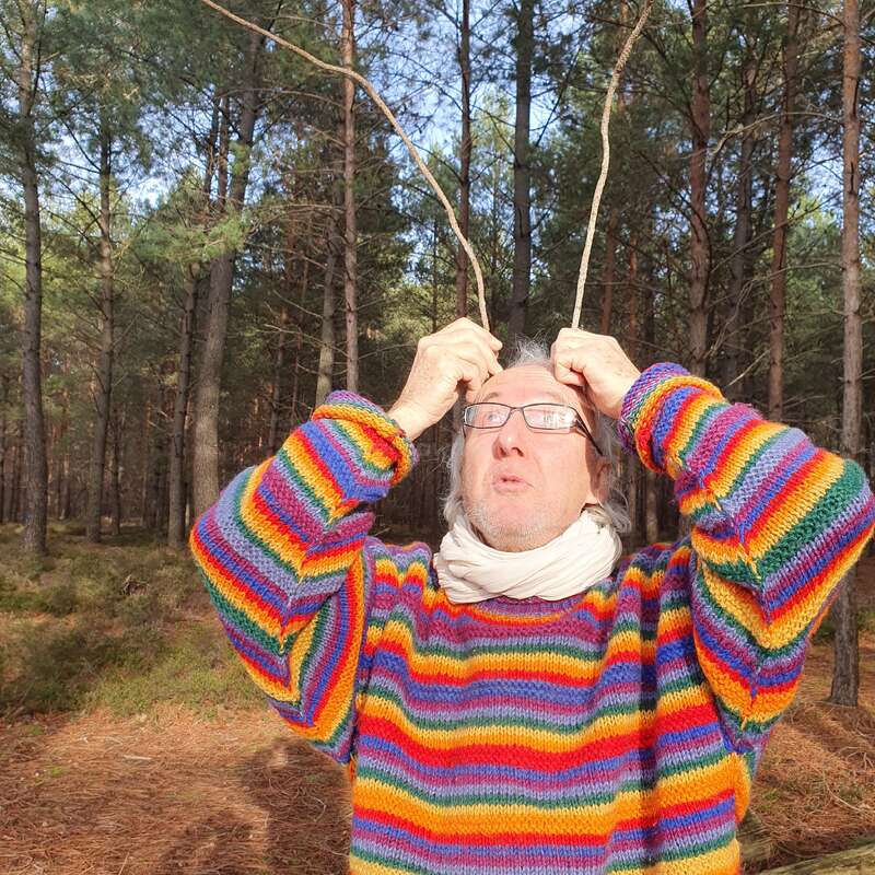 Um homem mais velho, com cabelos brancos e óculos, está em uma área arborizada, vestindo um suéter listrado e segurando duas varas acima da cabeça como chifres.