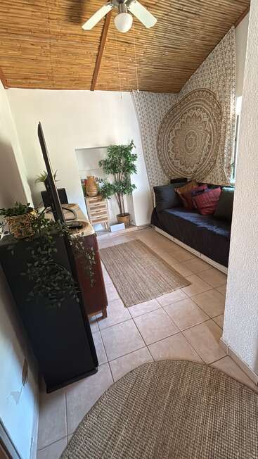 Esta acogedora habitación cuenta con un techo de bambú, suelo de baldosas, tapices de estilo bohemio, un diván negro con cojines, plantas, una alfombra estrecha, un ventilador y muebles de madera con detalles decorativos.