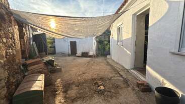 Un patio rústico con el suelo agrietado, a la sombra de un gran toldo de tela. La leña, dos sillas, una mesita y una manguera de jardín contribuyen a crear un ambiente acogedor.