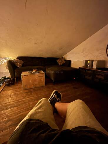 Um quarto aconchegante no sótão, com piso de madeira, iluminação suave, um sofá verde, almofadas decorativas, uma mesa rústica de madeira, um aparelho de som e alguém relaxando, com as pernas à mostra.
