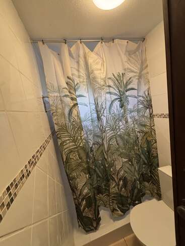 Um banheiro pequeno com paredes revestidas de azulejos brancos, uma faixa decorativa em mosaico, uma cortina de chuveiro com tema tropical com plantas verdes, um lustre e um vaso sanitário branco parcialmente visível em primeiro plano.