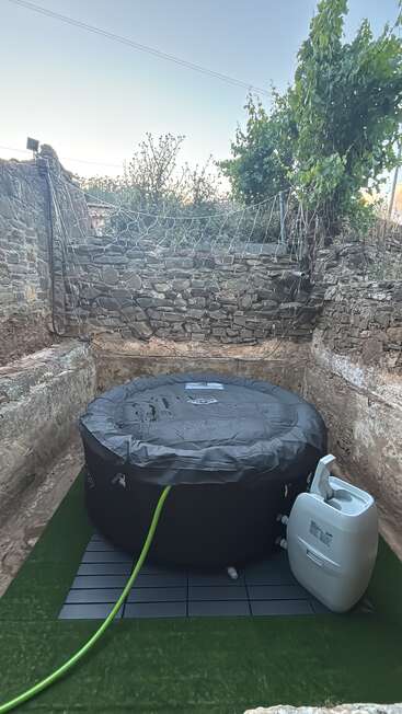 Se ha instalado un jacuzzi hinchable con cubierta al aire libre, entre muros rústicos de piedra, rodeado de vegetación, césped artificial y una red en la parte superior por motivos de seguridad.