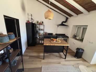 Esta acogedora cocina cuenta con una isla de madera con una cesta de fruta, estanterías abiertas con objetos de cerámica, electrodomésticos modernos, un suelo rústico y una lámpara colgante de mimbre.