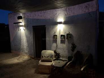 Una zona de descanso al aire libre al atardecer cuenta con dos sillones, una mesita, plantas en macetas y tres cuadros enmarcados. Hay una silla colocada de forma extraña en lo alto de la pared.