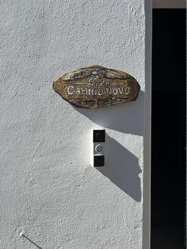Una pared blanca con textura luce un letrero rústico de madera en el que se lee «Lar do CAMINO NOVO» sobre un moderno videoportero, con la entrada proyectando una sombra cerca.