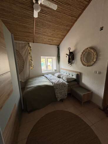Este acogedor dormitorio cuenta con una cama de matrimonio, una decoración de estilo boho, un techo de madera natural, un espejo redondo, plantas colgantes, una iluminación tenue, textiles estampados y un ambiente tranquilo y acogedor.