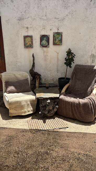 Dos cómodos sillones con mantas, una mesa rústica de piedra y madera, una planta en maceta, una figurita religiosa y tres cuadros enmarcados de santos colgados en una pared blanca desgastada.
