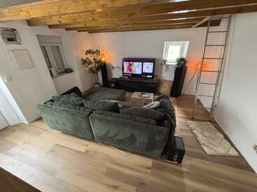 Un acogedor salón con suelos de madera, un sofá modular verde, un televisor, dos plantas en macetas, iluminación ambiental, vigas vistas, una alfombra, una ventana pequeña y una escalera.