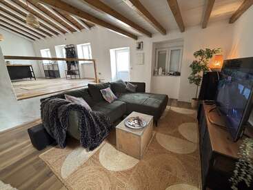 Este acogedor salón cuenta con un gran sofá modular verde, un televisor, vigas de madera natural, una iluminación tenue, plantas de interior y un diseño moderno de dos niveles y concepto abierto.