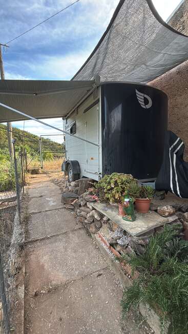 Hay una caravana aparcada junto a un muro de piedra, bajo un toldo. Unas plantas suculentas y un pato de cerámica decoran un pequeño jardín bordeado de piedra, rodeado por una valla rústica.