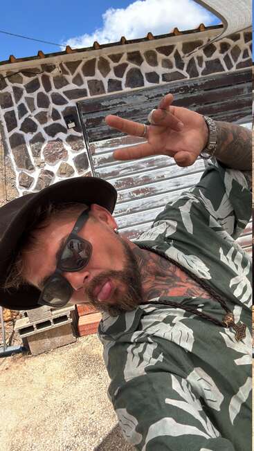 Un hombre barbudo, con gafas de sol y gorra, posa de forma divertida para hacerse un selfi, haciendo el signo de la paz, con un muro de piedra y un cielo azul a sus espaldas.