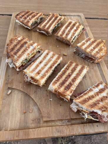 En esta imagen se ve una tabla de cortar de madera con ocho mitades de sándwich a la plancha, dispuestas de forma ordenada. Los sándwiches presentan marcas de la parrilla y contienen capas de verduras, queso y carne.