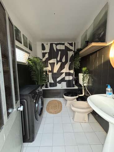 This modern bathroom features a washing machine, black-and-white decor, green plants, a patterned shower curtain, artwork, a sink, toilet, bidet, and woven rug.