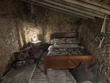 A cozy rustic bedroom with stone walls, a wooden bed, floral bedding, woven bench with colorful pillows, hanging plants, soft lighting, and a sloped ceiling.