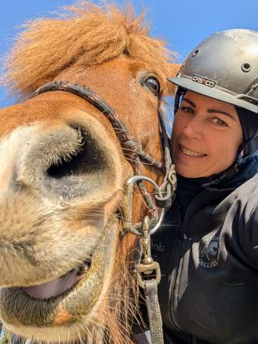 Eine Frau mit Helm lächelt neben einem braunen Pferd. Das Foto ist ein Selfie aus nächster Nähe. Beide wirken glücklich, vor dem Hintergrund eines strahlend blauen Himmels.
