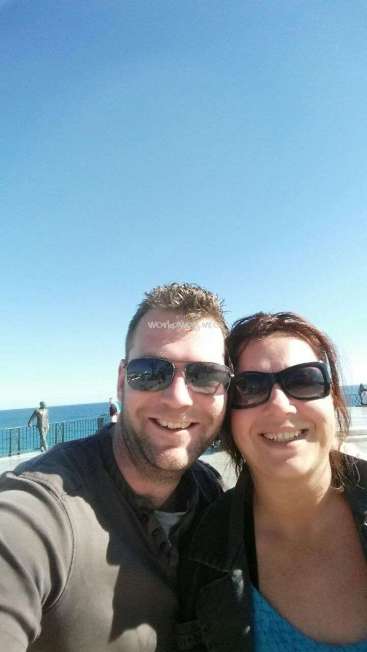 Das Bild zeigt ein Selfie von einem Mann und einer Frau, die vor einem Gewässer posieren, mit einem klaren blauen Himmel im Hintergrund.