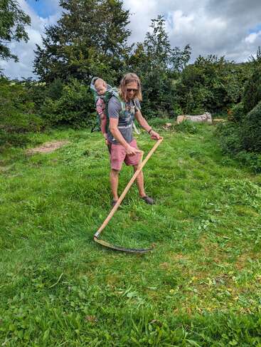 A man is mowing grass with a scythe while carrying a baby on his back in a carrier. Lush greenery surrounds them under a cloudy sky.