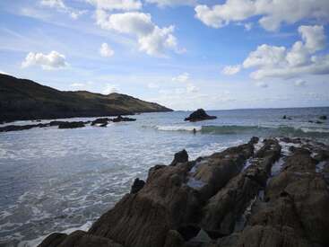 Rocky coastline meets gentle waves under a vibrant blue sky filled with fluffy white clouds. Hills rise in the distance; the landscape feels peaceful and serene.