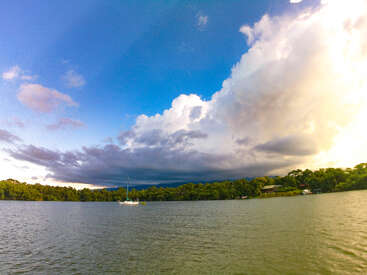 Ein ruhiger See mit einem einsamen Segelboot, umgeben von üppig grünen Bäumen. Der Himmel wird von dramatischen Wolken beherrscht, die von strahlendem Weiß in dunkles, stürmisches Grau übergehen.