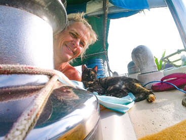 A smiling person with light hair poses on a boat next to a relaxing tortoiseshell cat, surrounded by nautical equipment, ropes, and bright sunlight.