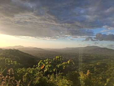 This image shows a breathtaking landscape of green hills and mountains under a dramatic, cloudy sky. Sunlight gently illuminates the scene, creating a peaceful, serene atmosphere.