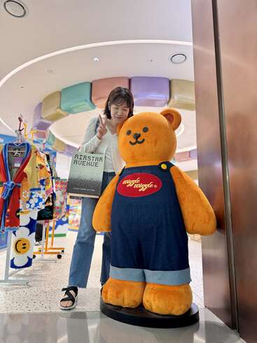 Uma mulher posa ao lado de uma grande estátua de ursinho de pelúcia laranja usando macacão jeans. Ela segura uma sacola de compras "AIRSTAR AVENUE" e faz um sinal de paz.