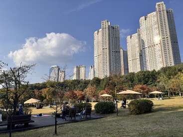 Pessoas relaxam em um parque com bancos, guarda-chuvas e árvores de outono. Prédios altos modernos se erguem ao fundo sob um céu azul com nuvens.