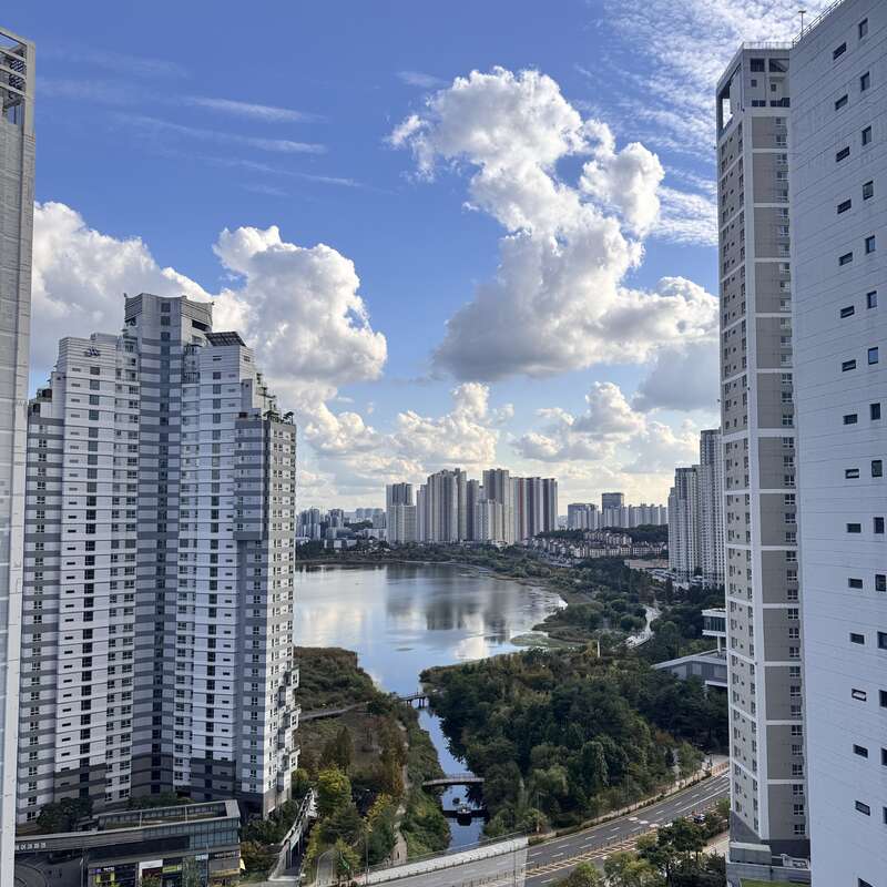Prédios de apartamentos altos e modernos emolduram uma bela paisagem urbana com um lago sereno, vegetação exuberante, estradas sinuosas e um céu azul vibrante pontilhado de nuvens fofas.