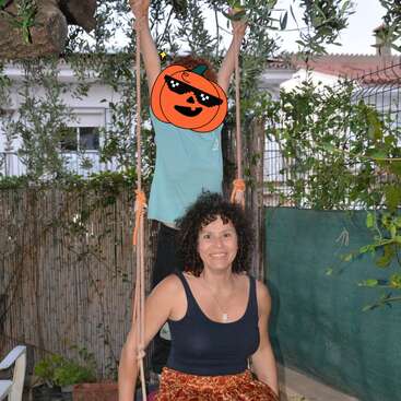 A woman with curly hair sits on a swing, smiling. Behind her, a child stands holding ropes, face covered with a cartoon pumpkin sticker, outdoors.