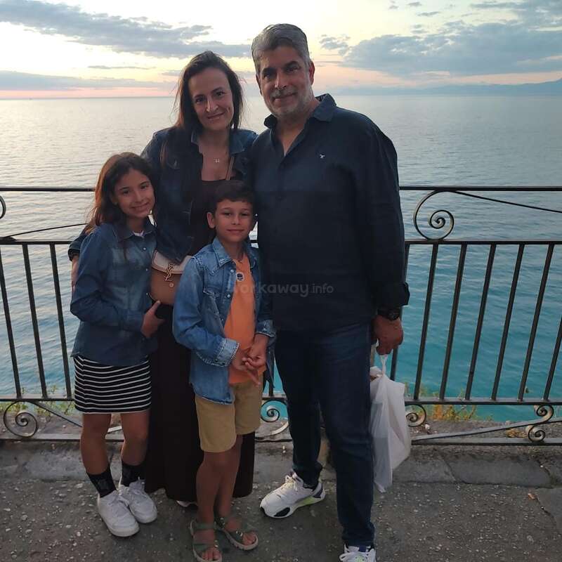 Eine vierköpfige Familie posiert bei Sonnenuntergang an einer Reling am Meer. Sie lächeln, sind leger gekleidet und genießen den schönen Blick auf das Meer bei bewölktem Himmel.
