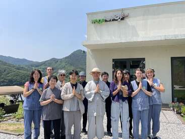 A group of people stand together outside a building with "vege" on the sign, smiling and making a prayer gesture, surrounded by mountains under clear skies.