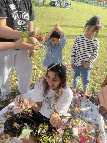 A joyful group enjoys outdoors on grass, surrounding a smiling woman with flowers. Children laugh and play, tossing petals, creating a festive, colorful, and lively atmosphere.