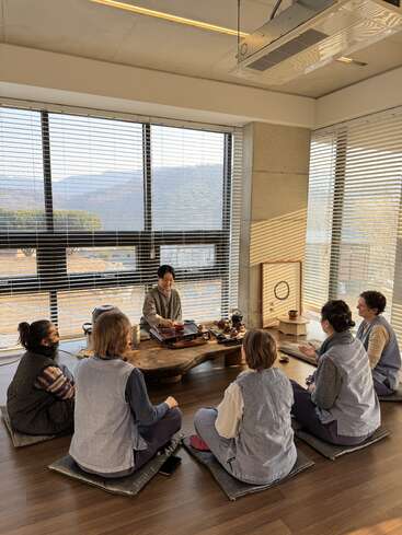 Sechs Personen sitzen auf Bodenkissen um einen niedrigen Holztisch herum und halten eine traditionelle Teezeremonie ab, während das Sonnenlicht durch große Fenster mit Blick auf die Berge fällt.