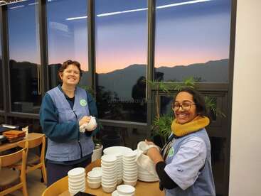 Two smiling women are drying dishes inside a room with large windows. A beautiful sunset and mountains can be seen outside. They seem cheerful and content.