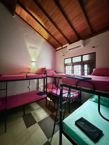 This image shows a hostel room with metal bunk beds, colorful bedding, wooden ceiling, air conditioner, window, and ambient wall lighting, creating a cozy shared living space.