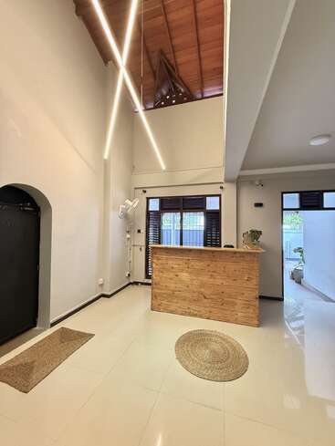 This image shows a modern, minimalist reception area with a wooden desk, high ceiling, striking diagonal lights, white tiled floors, plants, and natural light through windows.