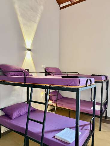 The image shows a clean dormitory room with two black metal bunk beds, neatly made with purple sheets, pillows, and white folded towels. Warm wall light.