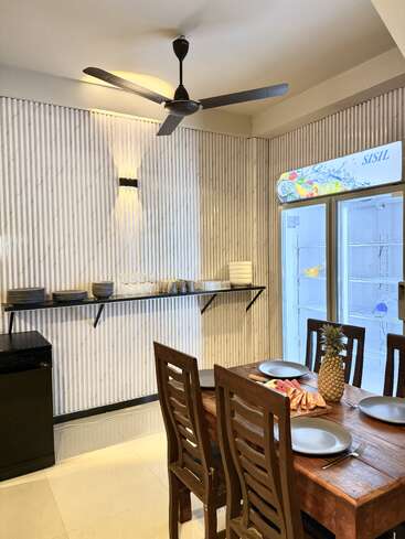A modern dining area with a wooden table set for four, featuring black plates and fresh fruit. Shelves hold dishes; a ceiling fan and fridge are present.