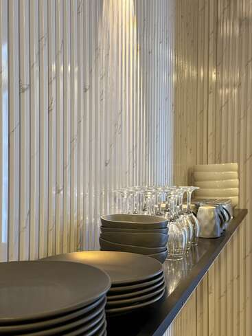 A sleek, modern shelf holds stacked dark plates, bowls, glassware, and white cups against a vertically striped, marble-patterned wall, reflecting warm ambient lighting elegantly.