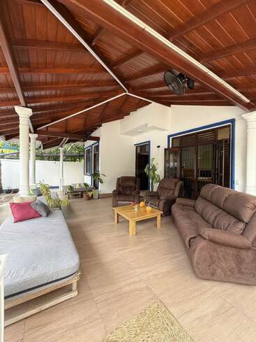 This image shows a spacious, covered patio with brown leather sofas, a wooden coffee table, large green plants, white pillars, and a wood-paneled ceiling. Peaceful ambiance.