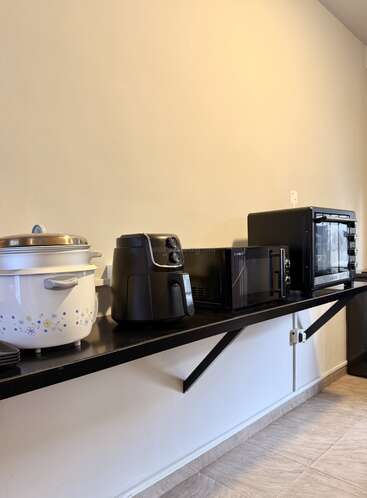A kitchen counter displays a rice cooker, air fryer, microwave, and oven. The appliances are neatly lined up on a black floating shelf against a beige wall.