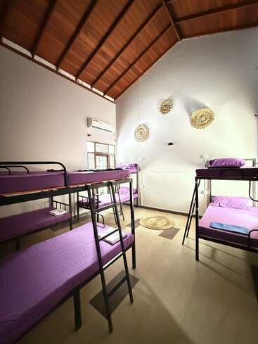 This image shows a clean, spacious dormitory room with four bunk beds featuring purple bedding. The room has a high wooden ceiling, air conditioning, and decorative wall pieces.