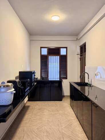 This image shows a modern, minimalist kitchen with black cabinets, a tiled floor, various appliances, a rice cooker, a sink, and a window with shutters.