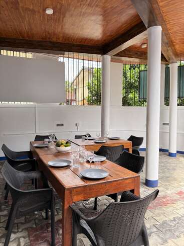 This image shows an outdoor dining area with a wooden table, black chairs, plates, glasses, and a fruit bowl, under a wooden roof with white pillars.